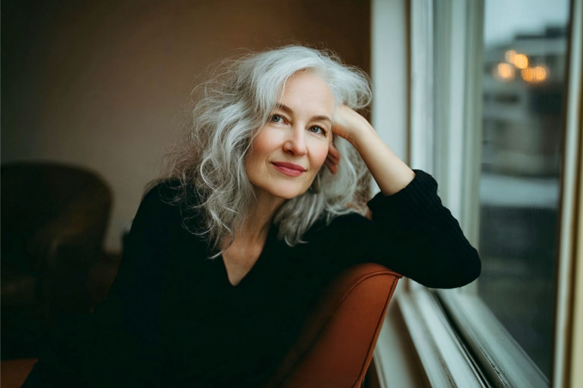 A woman with long gray hair and a black top sits by a window, resting her head on her hand and smiling softly. The background is softly lit and out of focus.