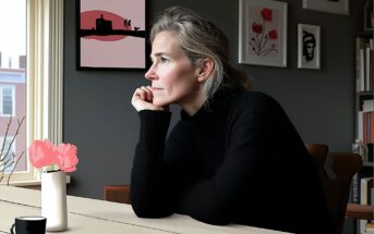A woman with gray hair, wearing a black sweater, sits at a table by a window, resting her chin on her hand and looking outside. A vase with pink flowers and framed art are visible in the softly lit room.