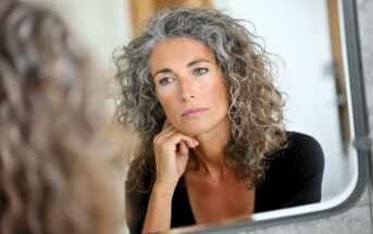 A woman with curly gray hair looks thoughtfully at her reflection in a mirror, resting her chin on her hand. She appears calm and introspective, wearing a black top in a softly lit room.