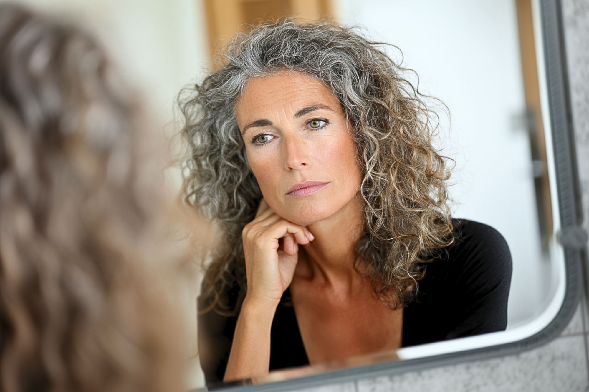 A woman with curly gray hair looks thoughtfully at her reflection in a mirror, resting her chin on her hand. She appears calm and introspective, wearing a black top in a softly lit room.