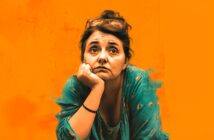 A woman with dark hair, wearing a teal top and glasses on her head, rests her chin on her hand and looks thoughtfully ahead, set against a bright orange background.