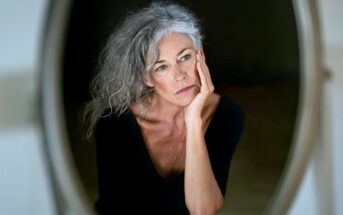 A woman with gray hair and a thoughtful expression rests her face on her hand, looking slightly to the side. She wears a black top and is reflected in a mirror against a dark background.