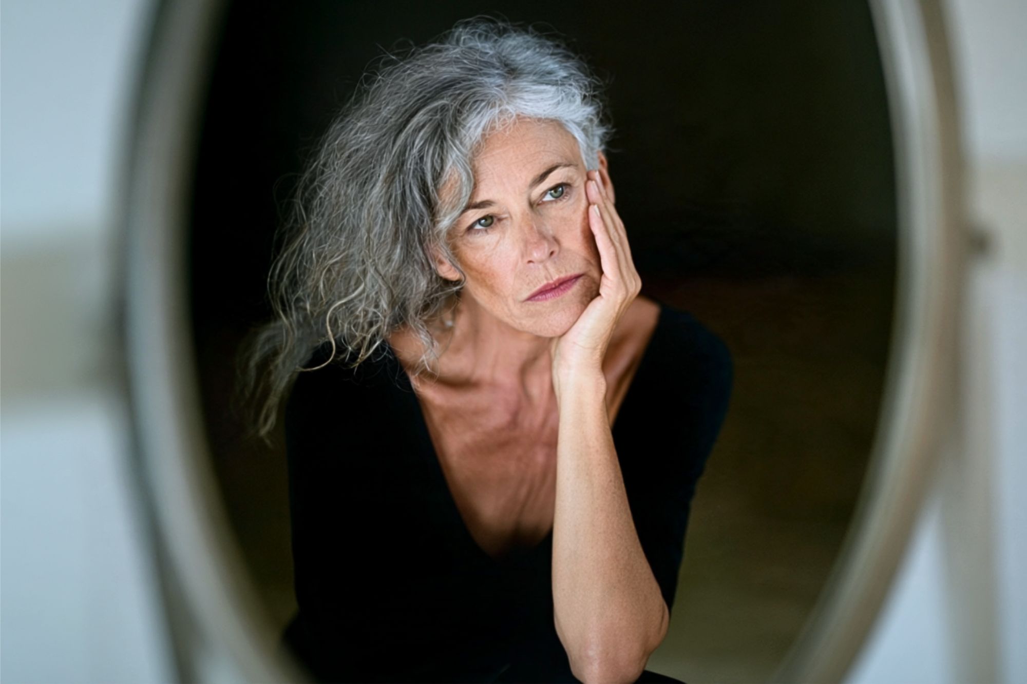 A woman with gray hair and a thoughtful expression rests her face on her hand, looking slightly to the side. She wears a black top and is reflected in a mirror against a dark background.