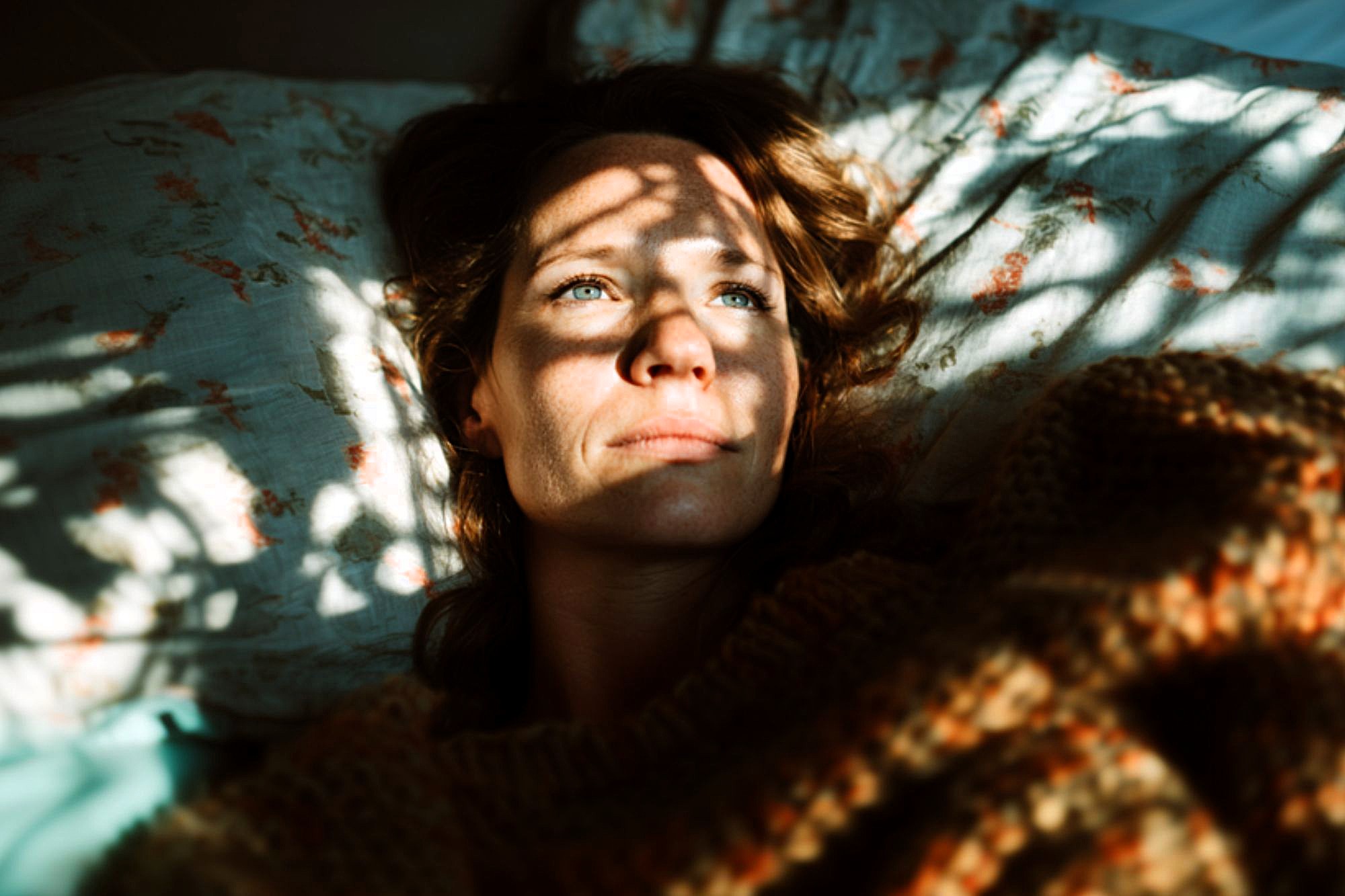 A woman lies on a bed, partially covered by a blanket, with sunlight and shadows from leaves cast across her face. She gazes upward with a calm, contemplative expression.