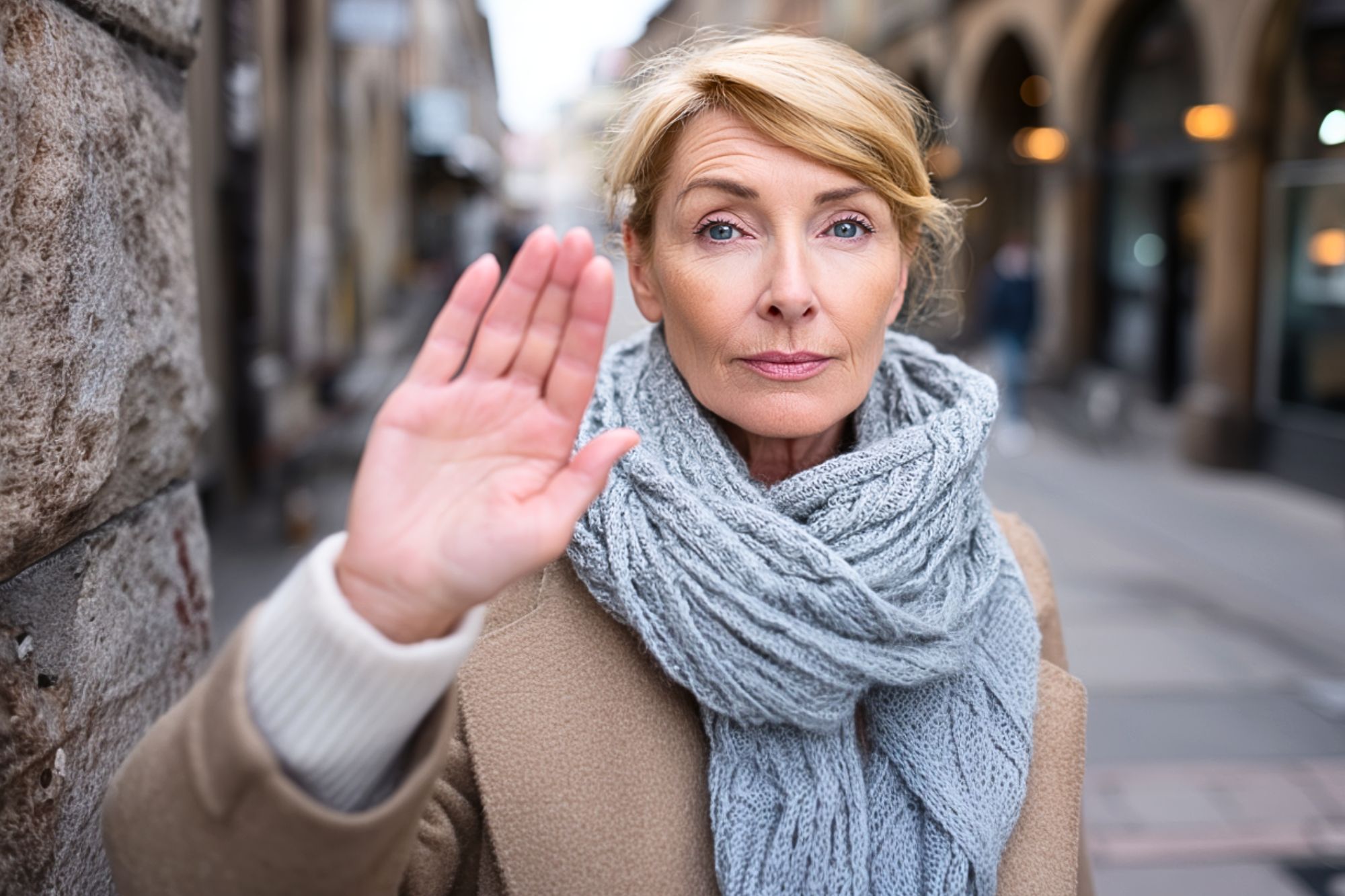A middle-aged woman with blonde hair stands outdoors in a city street, wearing a beige coat and a chunky gray scarf. She holds up her hand, palm forward, with a serious expression, as if signaling stop.