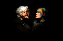 An older woman with white hair and glasses shouts at a younger woman with light hair, who looks away calmly. Both are in dark clothing, illuminated against a black background, with rainbow light across the younger woman’s face.
