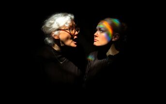An older woman with white hair and glasses shouts at a younger woman with light hair, who looks away calmly. Both are in dark clothing, illuminated against a black background, with rainbow light across the younger woman’s face.