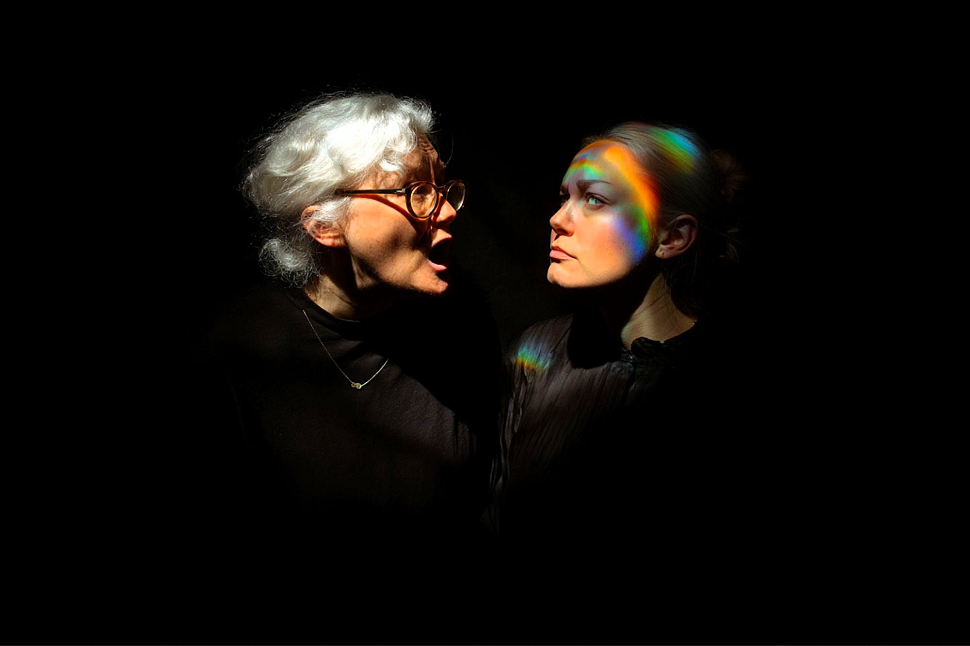 An older woman with white hair and glasses shouts at a younger woman with light hair, who looks away calmly. Both are in dark clothing, illuminated against a black background, with rainbow light across the younger woman’s face.
