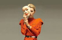 A woman in an orange dress holds a large, smiling mask close to her face, partially covering it, against a plain background. The mask is white with orange lips and eyebrows.