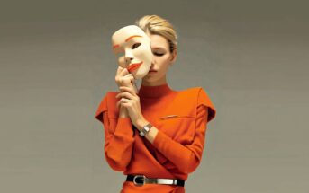 A woman in an orange dress holds a large, smiling mask close to her face, partially covering it, against a plain background. The mask is white with orange lips and eyebrows.