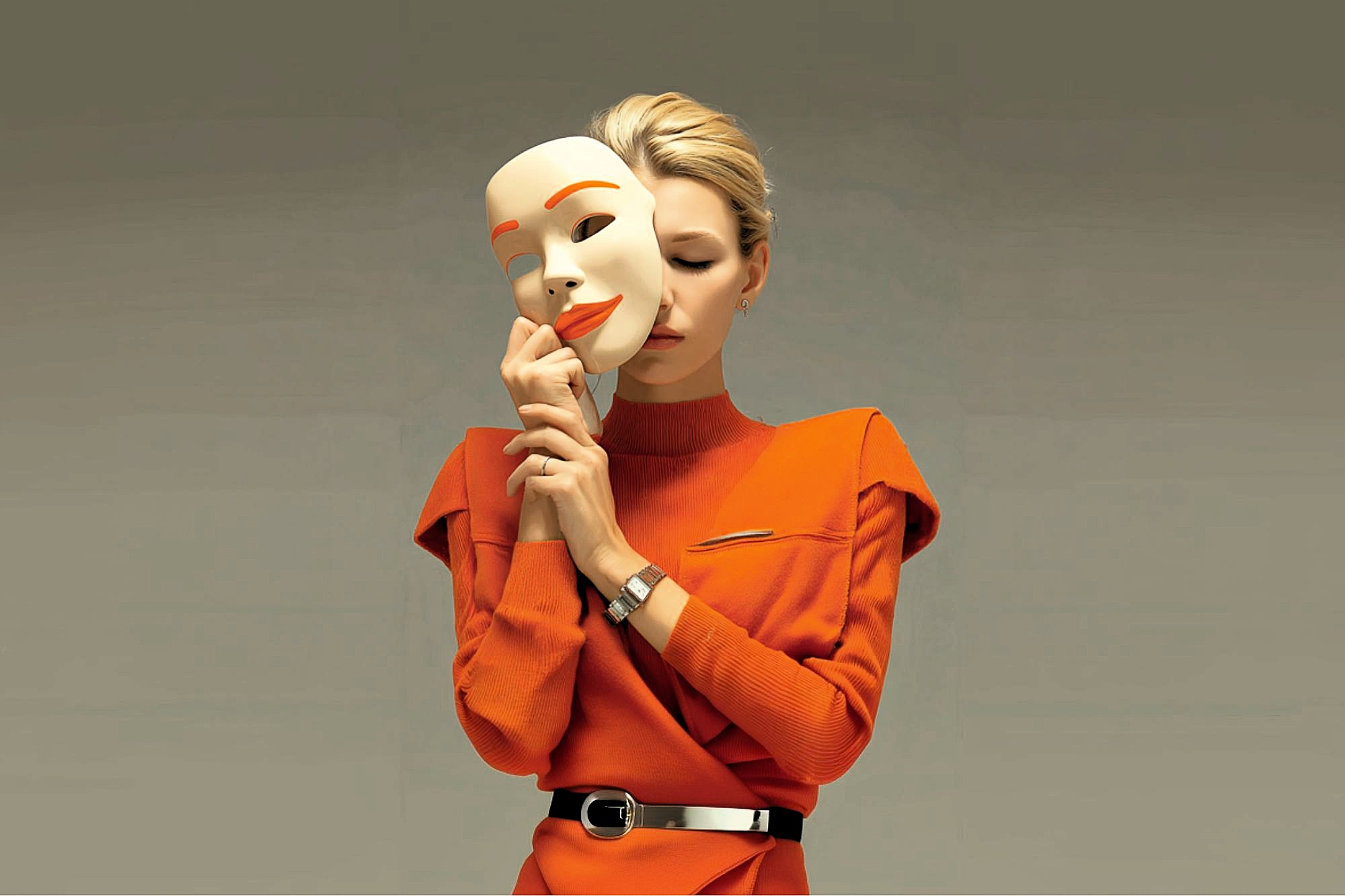 A woman in an orange dress holds a large, smiling mask close to her face, partially covering it, against a plain background. The mask is white with orange lips and eyebrows.