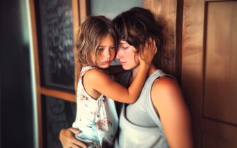 A young girl with short brown hair hugs an adult closely, both looking sad and thoughtful. The child wears a floral dress, and the adult wears a sleeveless gray shirt. They are sitting by a wooden wall indoors.