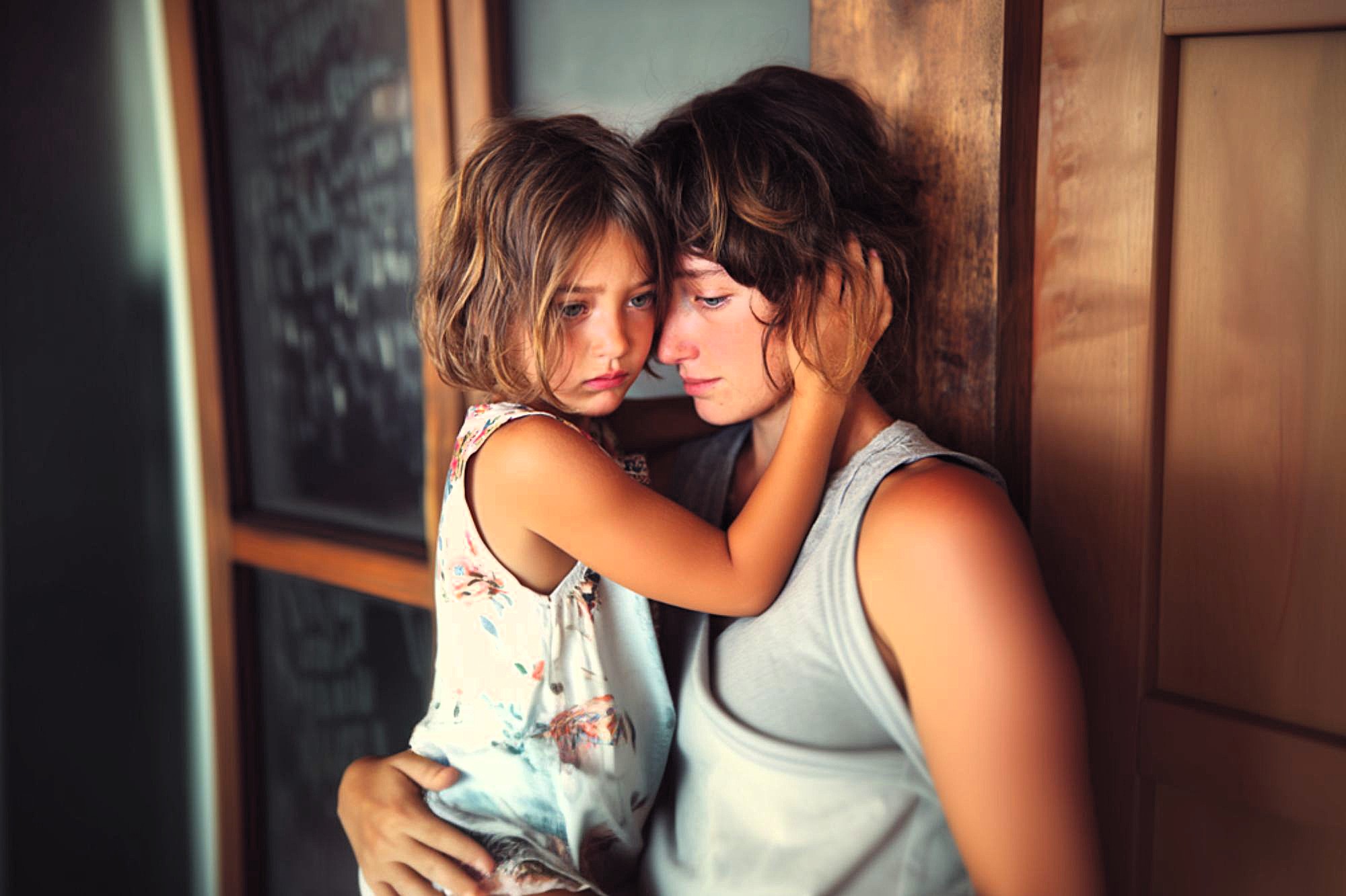 A young girl with short brown hair hugs an adult closely, both looking sad and thoughtful. The child wears a floral dress, and the adult wears a sleeveless gray shirt. They are sitting by a wooden wall indoors.