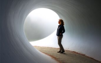 A person stands inside a large, curved tunnel, facing a bright circular opening that fills the space with soft light. The figure is silhouetted against the glowing entrance, creating a serene and contemplative scene.