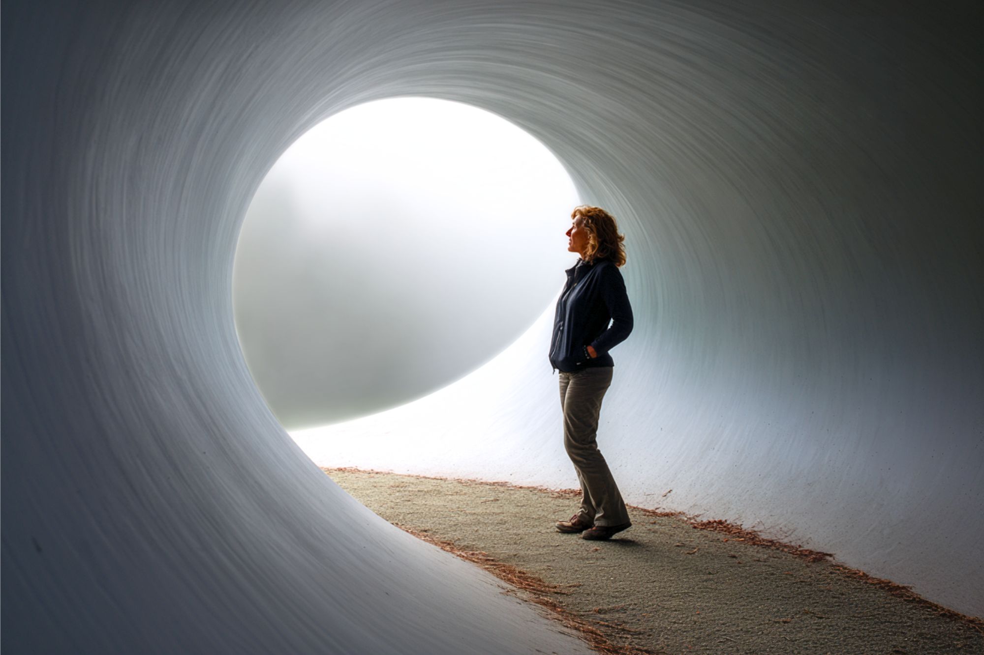 A person stands inside a large, curved tunnel, facing a bright circular opening that fills the space with soft light. The figure is silhouetted against the glowing entrance, creating a serene and contemplative scene.