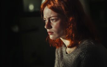 A young woman with red hair and a grey sweater looks pensive and serious, gazing into the distance with soft light illuminating her face against a dark background.