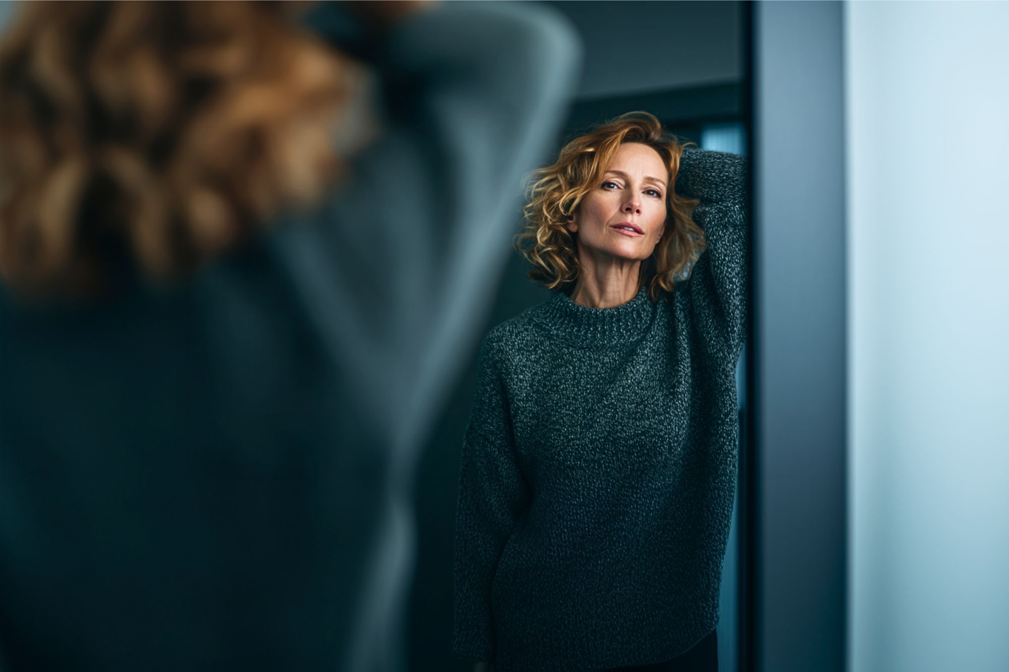 A woman with wavy blonde hair and a gray sweater stands by a mirror, gazing thoughtfully at her reflection with one arm resting on her head. The lighting is soft, creating a calm and introspective mood.