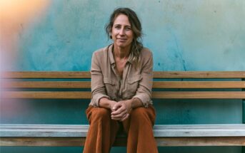 A woman with wavy hair sits on a wooden bench against a blue wall, smiling gently. She wears a beige shirt and rust-colored pants, with her hands clasped in front of her.