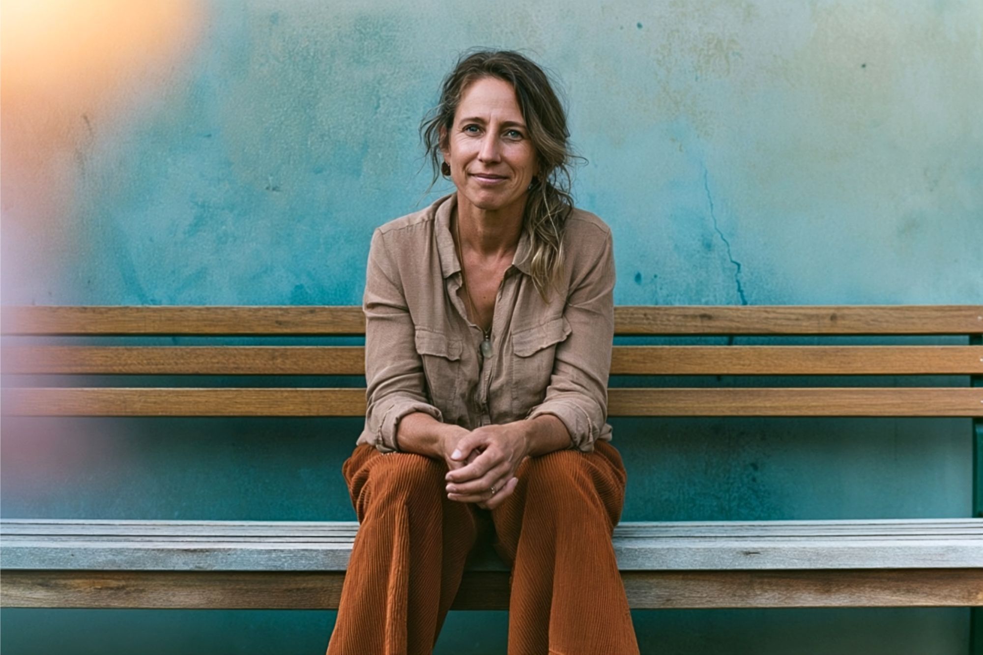 A woman with wavy hair sits on a wooden bench against a blue wall, smiling gently. She wears a beige shirt and rust-colored pants, with her hands clasped in front of her.