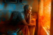 A person with short, light hair sits on a couch, gazing thoughtfully out a sunlit window. Warm orange and cool blue artistic splashes surround them, creating a dreamy, contemplative atmosphere.