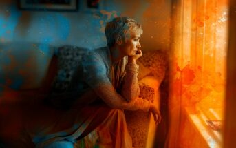 A person with short, light hair sits on a couch, gazing thoughtfully out a sunlit window. Warm orange and cool blue artistic splashes surround them, creating a dreamy, contemplative atmosphere.