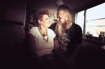 A smiling couple sits closely together on a couch in a warmly lit room, looking at each other affectionately. The woman wears a white top and chunky jewelry; the man has a long beard, tattoos, and ear gauges.