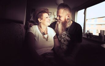 A smiling couple sits closely together on a couch in a warmly lit room, looking at each other affectionately. The woman wears a white top and chunky jewelry; the man has a long beard, tattoos, and ear gauges.