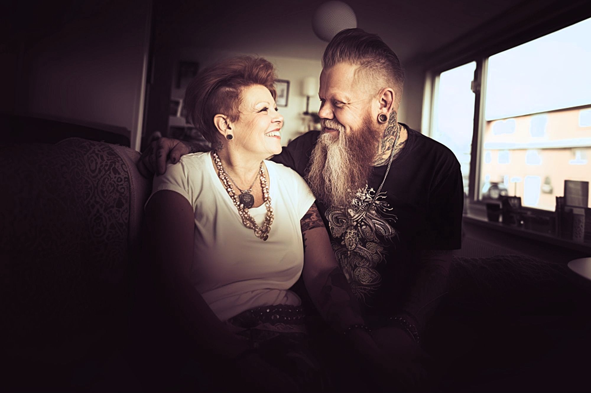 A smiling couple sits closely together on a couch in a warmly lit room, looking at each other affectionately. The woman wears a white top and chunky jewelry; the man has a long beard, tattoos, and ear gauges.