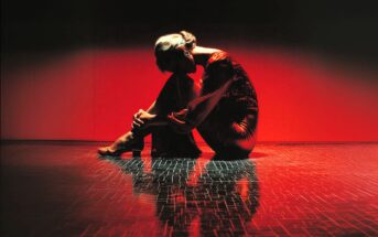 A person in a long dress sits on the floor with knees drawn close, head bent forward, and arms wrapped around their legs. Dramatic red lighting and dark shadows create a striking, emotional scene with a reflective floor.