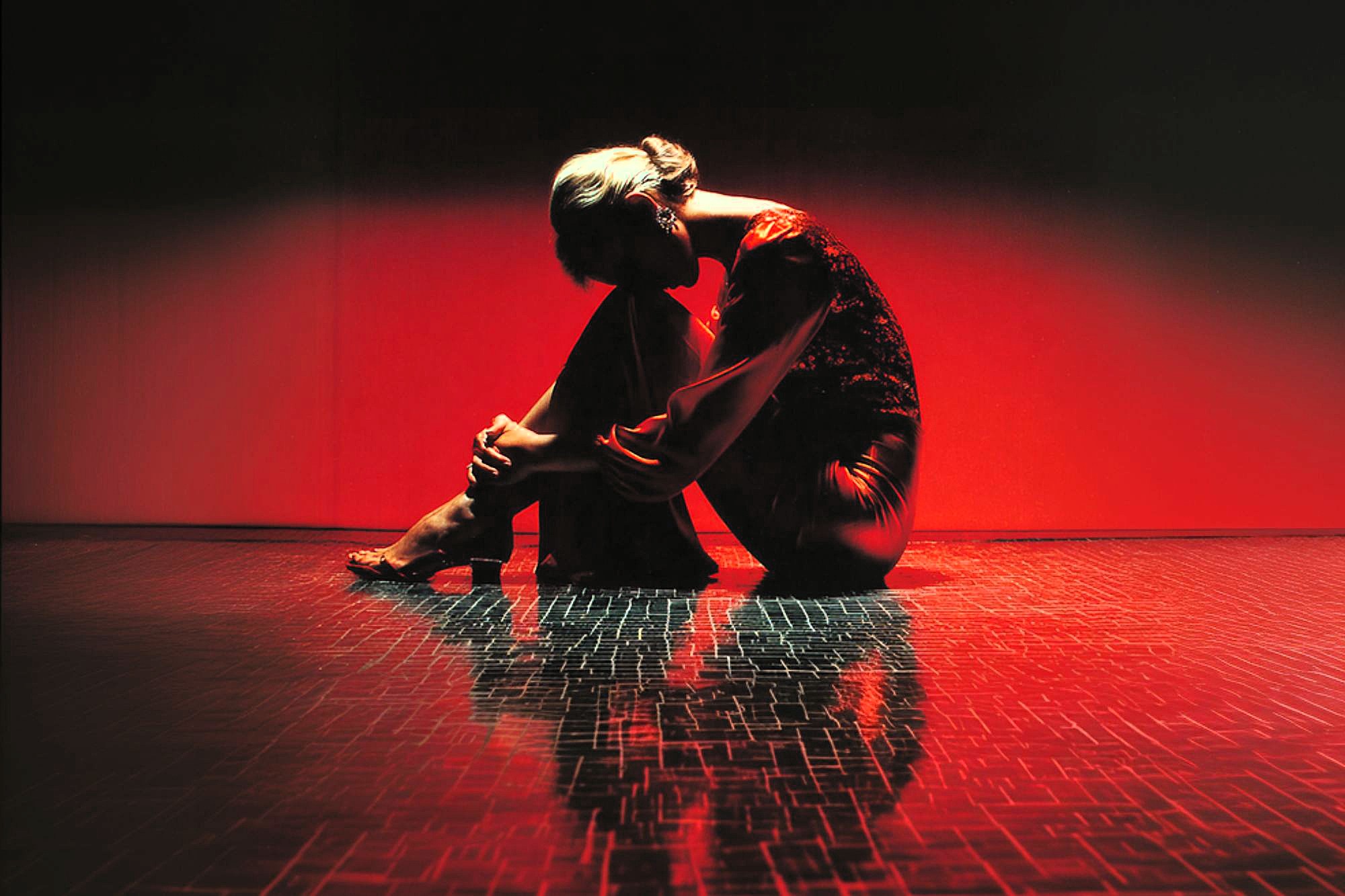 A person in a long dress sits on the floor with knees drawn close, head bent forward, and arms wrapped around their legs. Dramatic red lighting and dark shadows create a striking, emotional scene with a reflective floor.