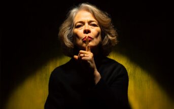 An older woman with gray hair and red lipstick holds her finger to her lips in a "shh" gesture, signaling for silence. She is wearing a black top and is illuminated by dramatic lighting against a yellow background.
