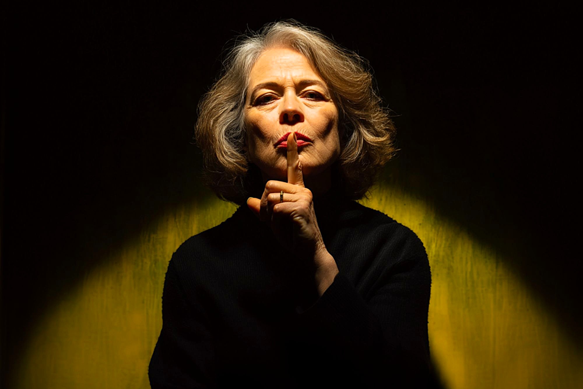 An older woman with gray hair and red lipstick holds her finger to her lips in a "shh" gesture, signaling for silence. She is wearing a black top and is illuminated by dramatic lighting against a yellow background.