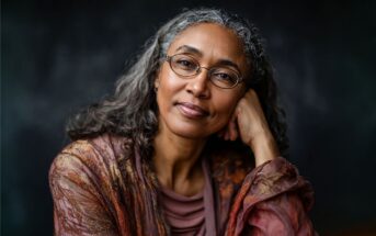 A woman with long gray hair and glasses, wearing a patterned shawl, sits with her head resting on her hand, looking calmly at the camera against a dark background.