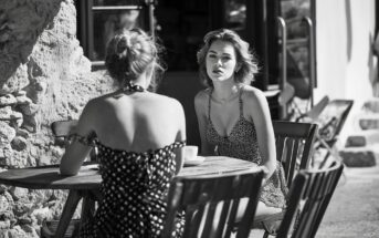 Two women in patterned summer dresses sit at a small outdoor café table, engaged in conversation. One woman faces the camera while the other has her back turned. The scene is in black and white.