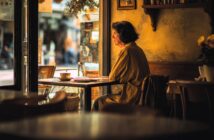 A woman with short dark hair sits alone at a small table by a window in a cozy café, wearing a tan dress and looking outside thoughtfully. Warm, soft light fills the quiet, rustic space.