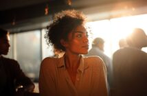 A woman with curly hair looks thoughtfully to the side, bathed in warm sunlight from a window behind her. She wears a light blouse and sits in a softly lit, indoor setting with blurred people in the background.