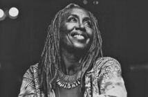 Black and white photo of a smiling woman with long dreadlocks, wearing a patterned jacket and a large, spiked necklace, looking slightly upward against a dark background.