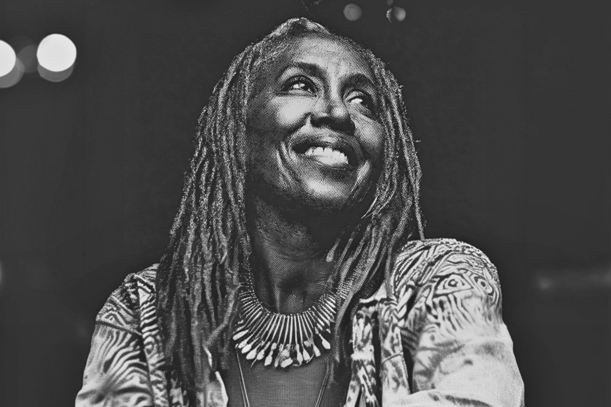 Black and white photo of a smiling woman with long dreadlocks, wearing a patterned jacket and a large, spiked necklace, looking slightly upward against a dark background.