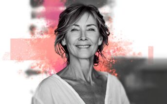 Smiling older woman with short hair, wearing a white top, stands against a blurred background with pink artistic splashes behind her.