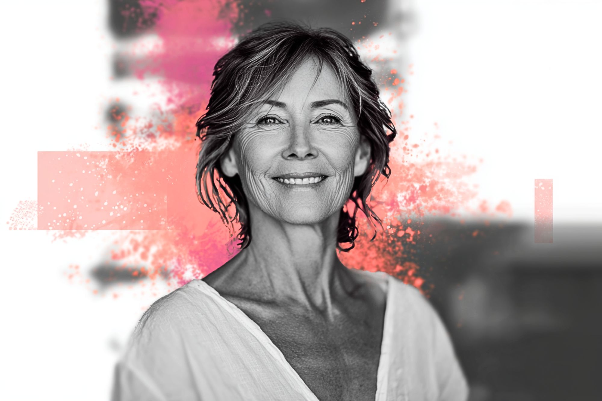 Smiling older woman with short hair, wearing a white top, stands against a blurred background with pink artistic splashes behind her.