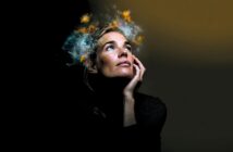 A woman gazes upward with her hand on her cheek, while bright, colorful clouds and sparks appear to emanate from her head against a dark background, symbolizing imagination or creativity.