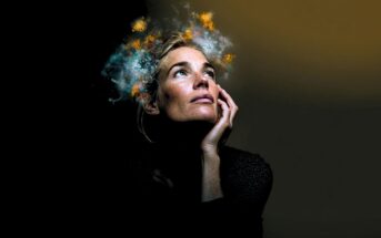 A woman gazes upward with her hand on her cheek, while bright, colorful clouds and sparks appear to emanate from her head against a dark background, symbolizing imagination or creativity.