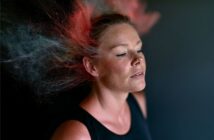 A woman with closed eyes stands against a dark background as clouds of red and blue powder explode around her head, creating a dynamic, colorful effect.
