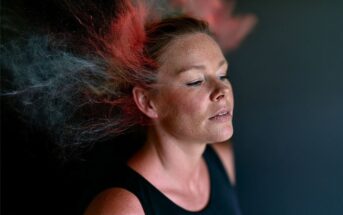 A woman with closed eyes stands against a dark background as clouds of red and blue powder explode around her head, creating a dynamic, colorful effect.