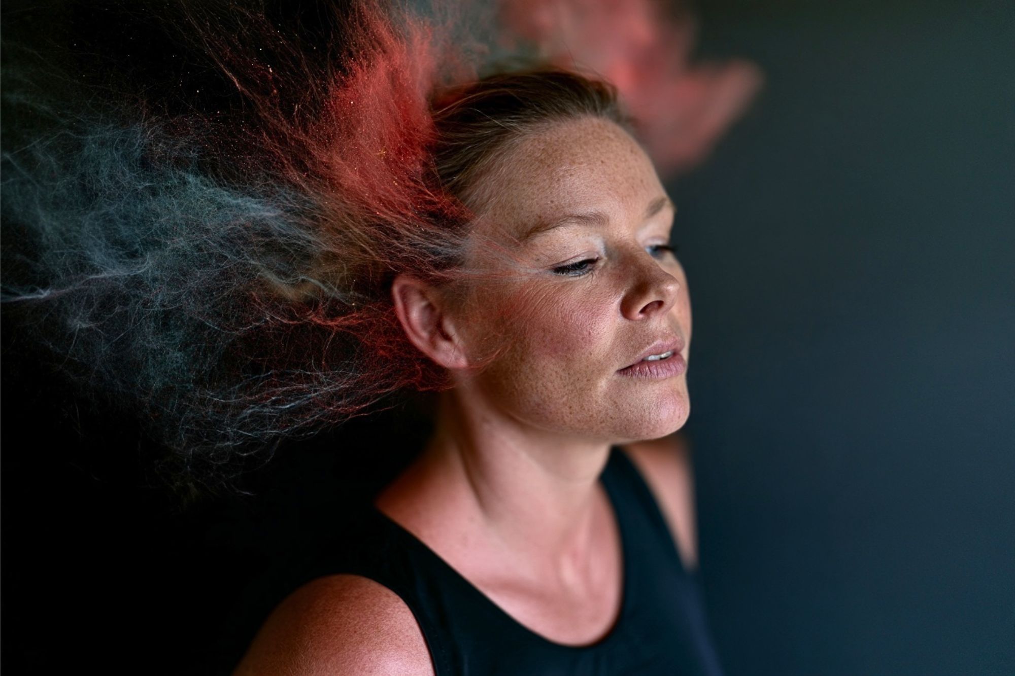A woman with closed eyes stands against a dark background as clouds of red and blue powder explode around her head, creating a dynamic, colorful effect.