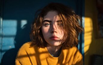 A young person with short, wavy brown hair and green eyes wears a yellow sweater, looking into the camera as sunlight casts shadows and lines across their face and blue background.