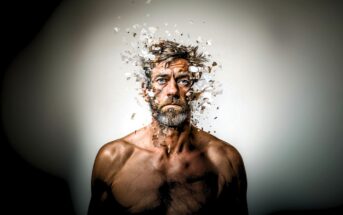 A shirtless man with a somber expression appears to have parts of his head and face breaking apart into small, scattered fragments, giving a surreal, dissolving effect against a plain background.