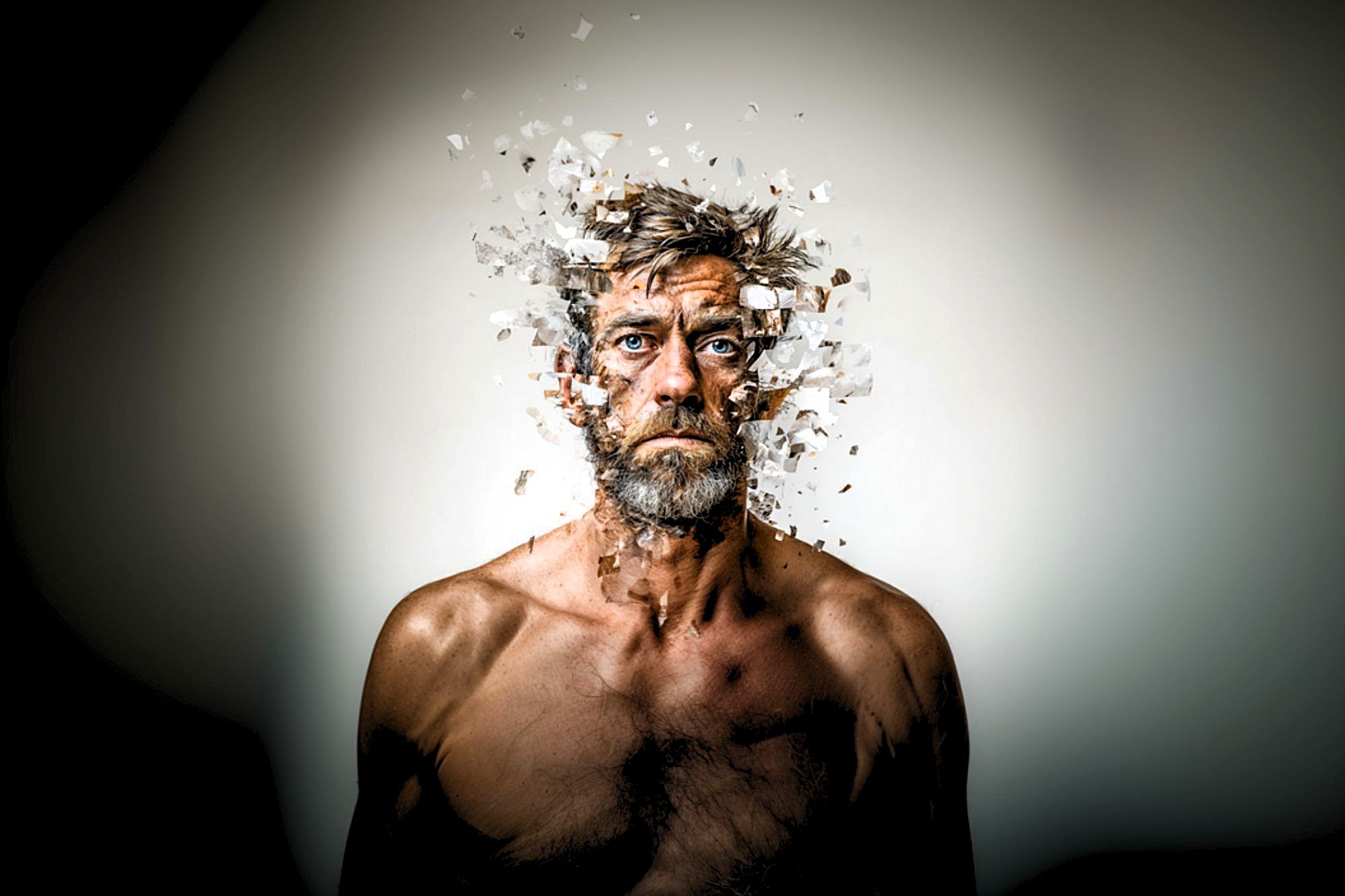 A shirtless man with a somber expression appears to have parts of his head and face breaking apart into small, scattered fragments, giving a surreal, dissolving effect against a plain background.