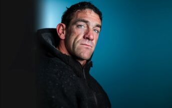A man with short, dark, curly hair and blue eyes wears a black textured jacket, gazing seriously at the camera against a smooth blue background.