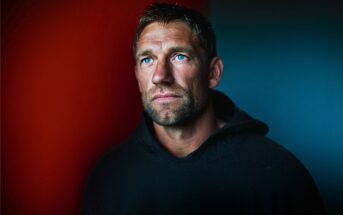 A man with short brown hair, blue eyes, and stubble gazes upward. He wears a black hoodie. The background is split between a red and blue gradient, creating a dramatic effect.